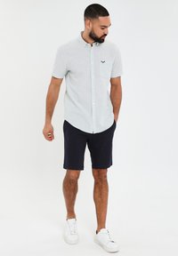 Camisa de botones de manga corta verde claro con un bolsillo en el pecho y un logo, combinada con pantalones cortos azul marino y zapatillas blancas. Textura suave.