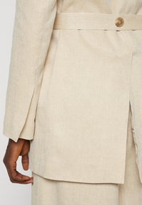 Beige Leinenjacke mit einer weichen Textur, langen Ärmeln und einem seitlichen Klappdetail mit einem passenden Knopfverschluss.