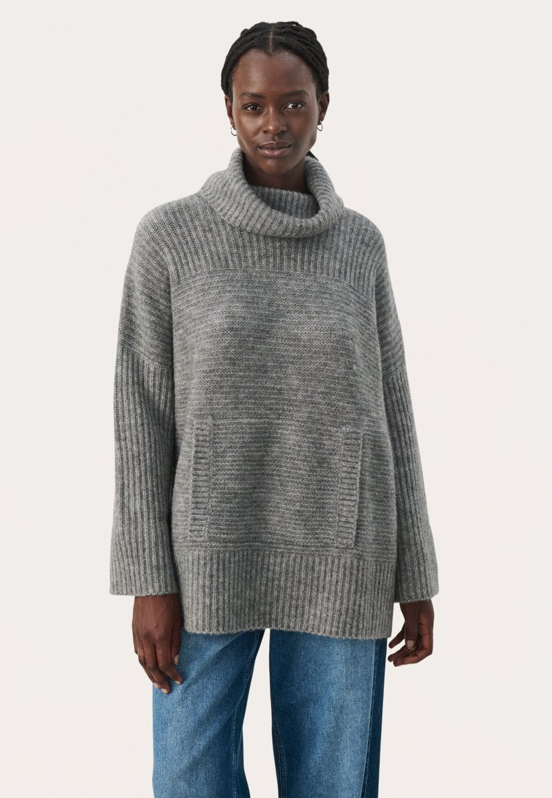 Gray knitted sweater with a high cowl neck, ribbed accents, two front pockets, and a relaxed fit, paired with blue jeans.