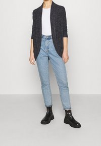 Cardigan lungo grigio scuro su una maglietta bianca, abbinato a jeans a vita alta azzurri e stivaletti neri. Tessuto morbido e vestibilità rilassata.