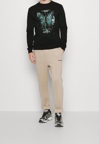 Felpa nera con design grafico di farfalla, pantaloni in pile beige con logo "PEGAADO" e sneakers nere con dettagli argentati.