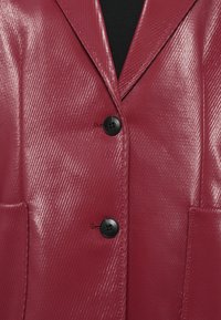 Blazer cremisi realizzato in materiale lucido con superficie texture, bottoni doppi, rever a punta e due tasche anteriori.