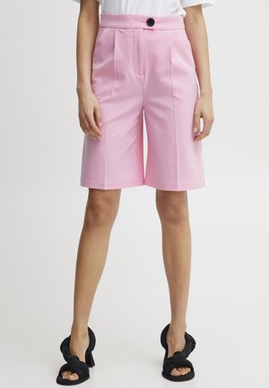 Bermuda shorts rose ajustés avec des plis et une fermeture à bouton, présentant une coupe droite et une texture lisse. Sandales noires à nœud.