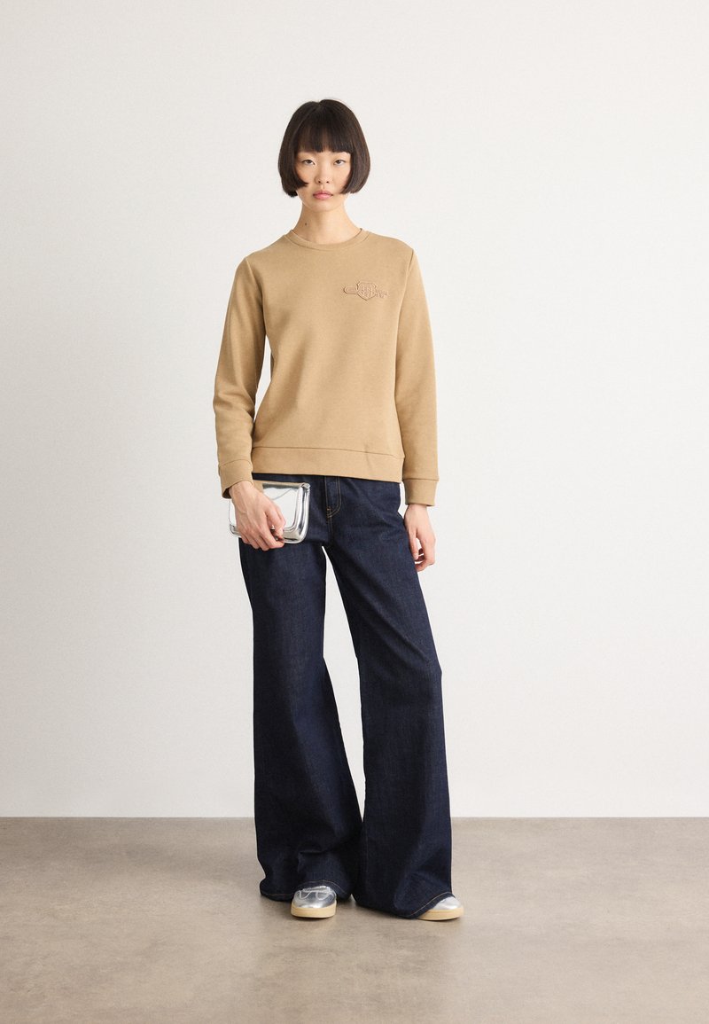 Frau mit kurzem Bob-Haarschnitt, die einen beigen Sweatshirt, weite dunkelblaue Jeans, silberne Sneakers trägt und eine kleine silberne Clutch vor einem einfarbigen Hintergrund hält.