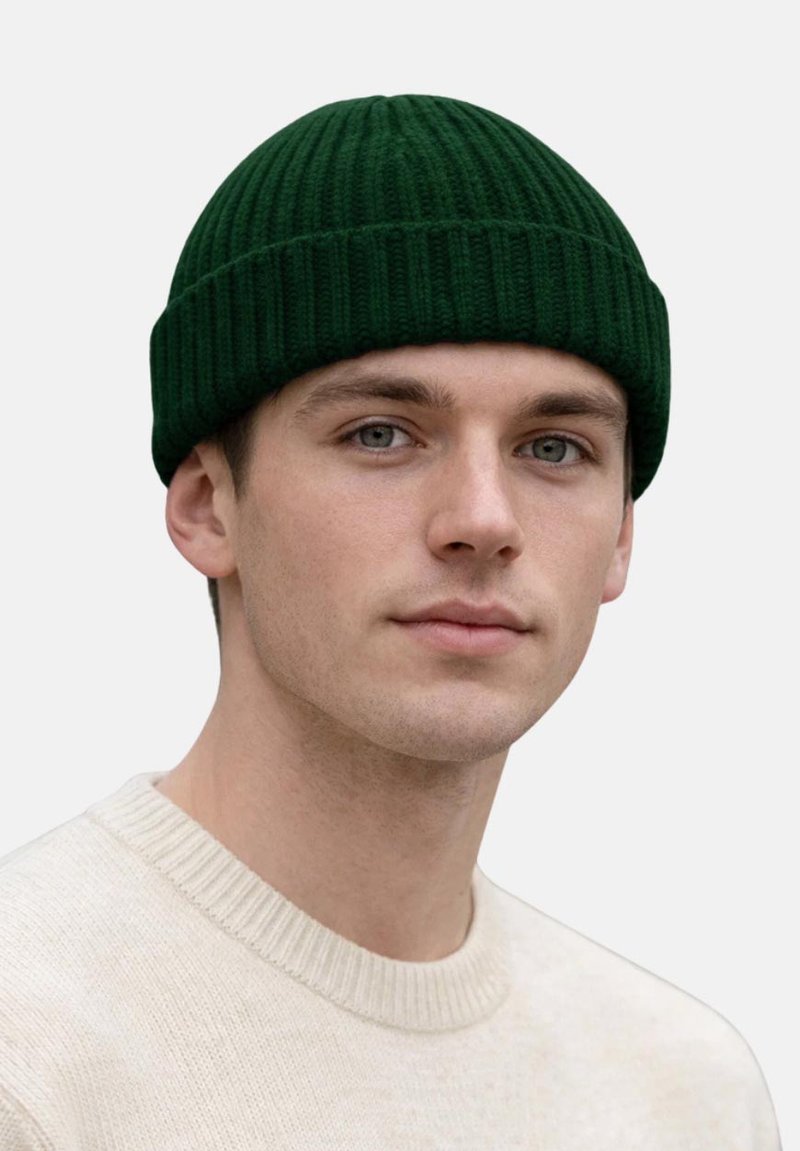 Junger Mann mit heller Haut und blauen Augen, der eine dunkelgrüne gerippte Strickmütze und einen cremefarbenen Pullover vor einfachem Hintergrund trägt.