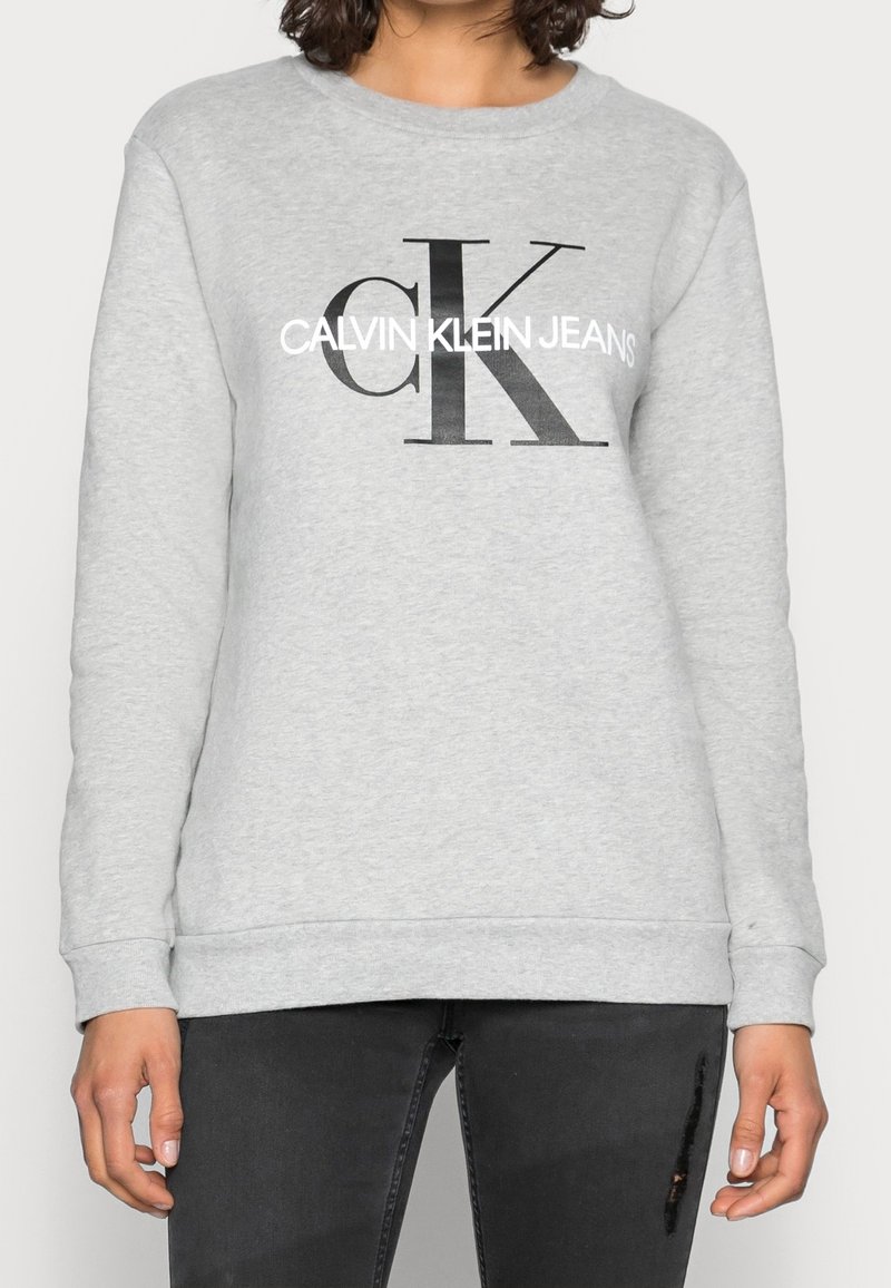 Personne portant un sweat-shirt gris clair avec un grand "cK" noir et le texte blanc "Calvin Klein Jeans" sur le devant, associé à un jean noir déchiré.