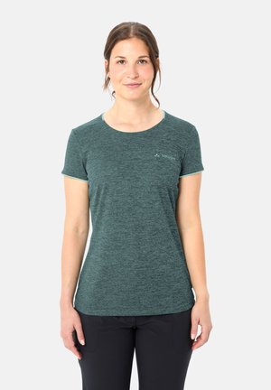 Junge Frau steht, trägt ein türkisfarbenes Vaude T-Shirt mit kurzen Ärmeln und schwarze Hose, schaut nach vorne mit neutralem Ausdruck.