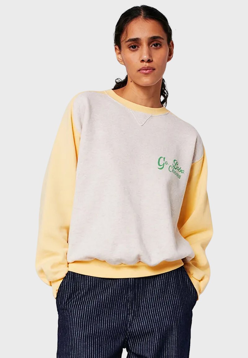 Giovane donna con capelli ricci scuri che indossa una felpa morbida gialla e bianca con testo verde e pantaloni scuri a righe.