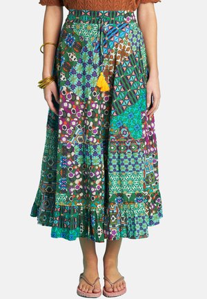 Woman wearing a colorful patchwork midi skirt with geometric patterns, tassel tie, bangles on left wrist, and flip-flop sandals, standing.