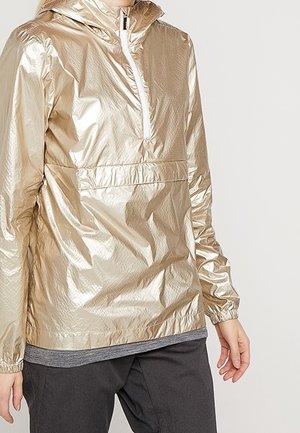 Persoon die een glanzende gouden windjack met capuchon draagt met een witte rits aan de voorkant en elastische manchetten, gecombineerd met een donkere broek en een grijs shirt eronder.