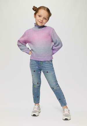 Paarse ombre gebreide trui met hoge kraag, gecombineerd met lichtblauwe jeans met kleurrijke vlinderborduursels en grijze sneakers.