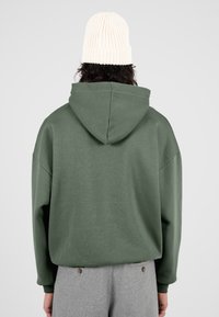 Persona con sudadera verde y gorro de punto blanco, vista desde atrás, de pie contra un fondo gris claro liso.