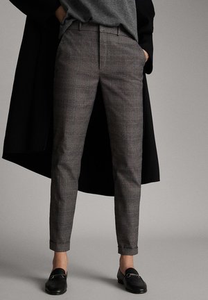 Osoba w szarych, wąskich spodniach w kratę, czarnych mokasynach i długim czarnym płaszczu, z rękami w kieszeniach.