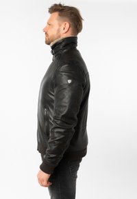 Chaqueta de cuero negra con un cuello alto, puños y dobladillo acanalados, que presenta un bolsillo lateral y un pequeño emblema en el hombro izquierdo.