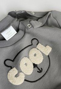 Felpa grigia con design di faccia di orso in peluche, caratterizzata dalla parola "cool" in tessuto beige testurizzato, chiusure a scatto in metallo sulla spalla.