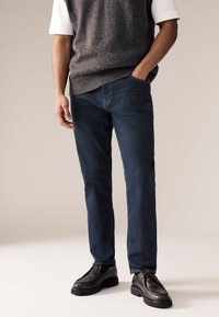 Tummansiniset farkut, suora malli, hienovaraisilla ompeluyksityiskohdilla ja sivutaskuilla, yhdistettynä harmaaseen neuleliiviin ja mustiin kenkiin.