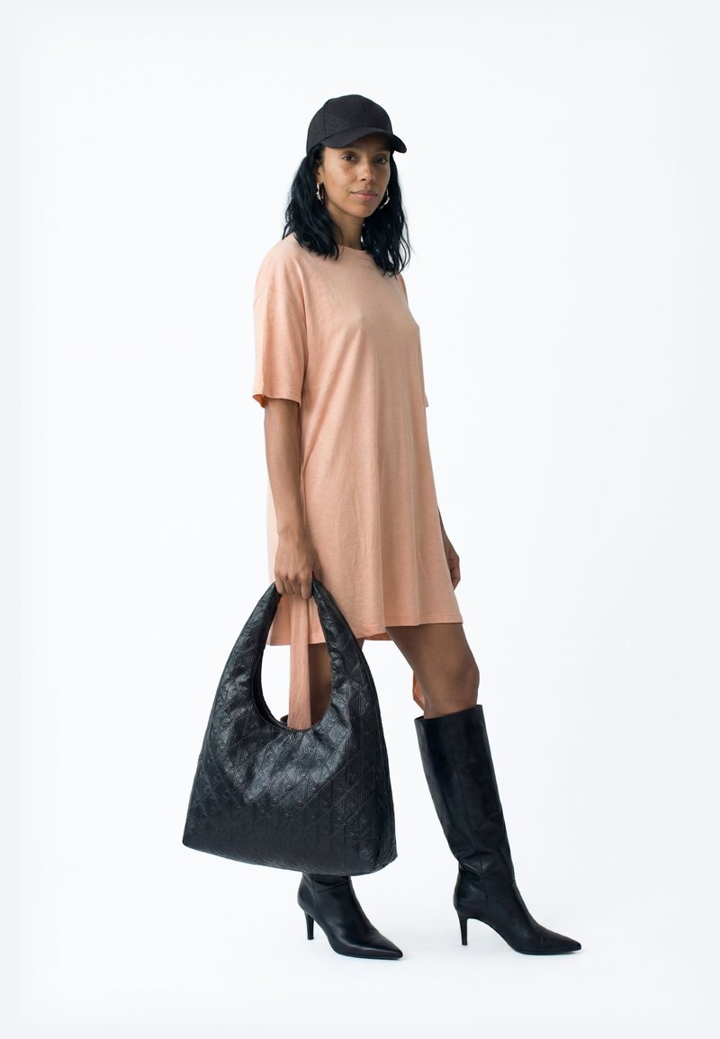 Mujer con un vestido camiseta oversize color melocotón, gorra negra, botas negras de tacón hasta la rodilla y sosteniendo un bolso grande negro con textura.