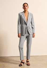 Grijs op maat gemaakt pak met een diep V-hals jasje, slim-fit broek en zwarte bandjeshakken. Glad materiaal en scherpe revers.