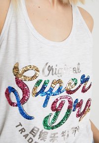 Camiseta sin mangas gris que presenta texto en lentejuelas multicolores "Original Super Dry" en varios colores llamativos. Textura de tela suave, diseño casual.