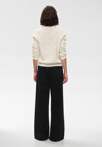 Femme aux cheveux bruns mi-longs, vêtue d’un pull en maille couleur crème et d’un pantalon noir à jambes larges, debout, de dos, sur un fond uni.