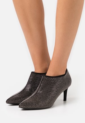 Black pointed-toe ankle boots with high heels, covered in small reflective studs, worn on bare feet against a white background.