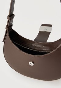 Καφέ δερμάτινη τσάντα χιαστί με ασημί μαγνητικό κούμπωμα, ρυθμιζόμενο ιμάντα και ορατή εσωτερική τσέπη με φερμουάρ, πάνω σε λευκό φόντο.