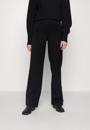 Person iført sorte bukser med lige ben, sort sweater og sorte ankelstøvler, stående mod en ensartet lys baggrund.