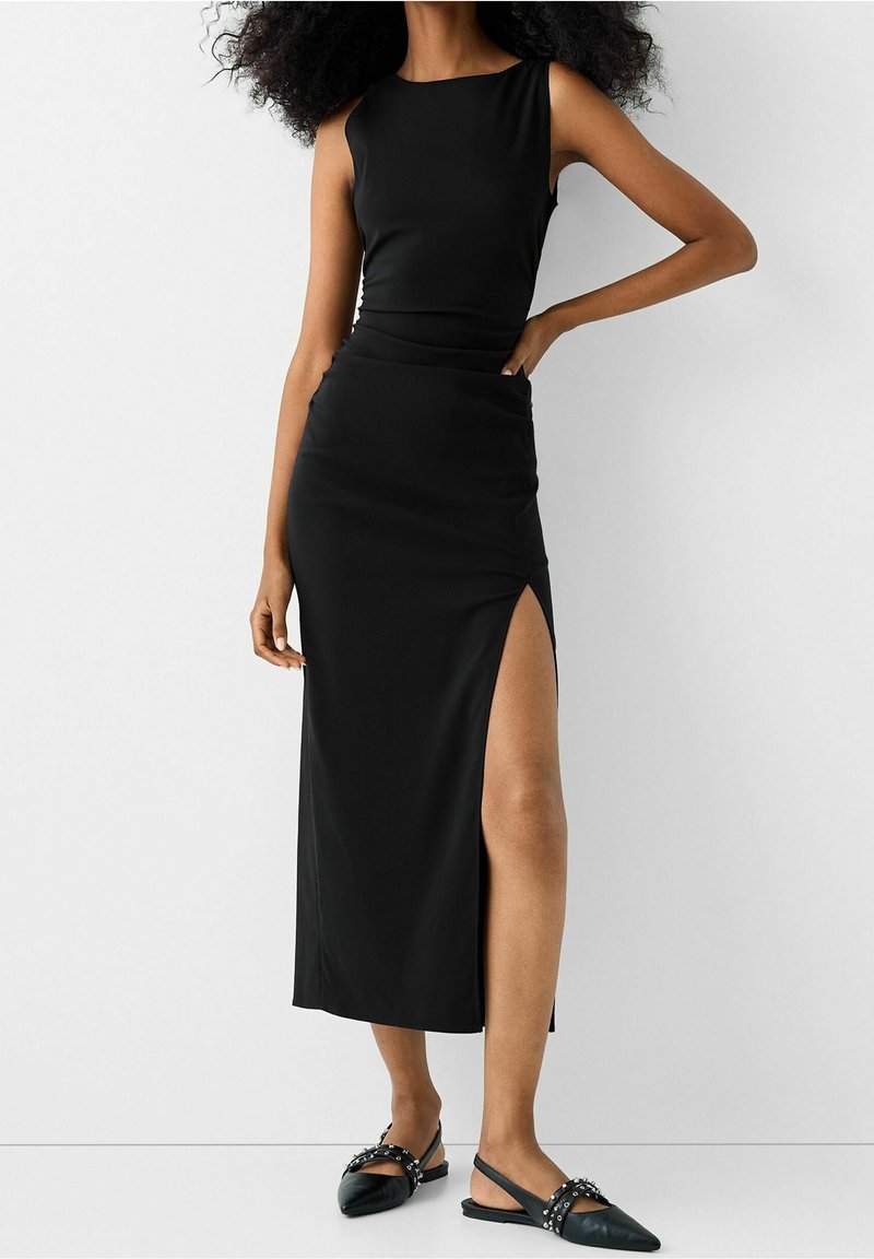 Abito nero senza maniche con un corpino aderente e una fessura alta fino alla coscia. Tessuto morbido, design semplice, abbinato a scarpe slip-on nere con borchie.