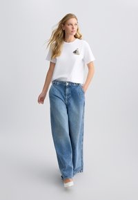 Hvid bomulds t-shirt med et mønstret sejlskib design på brystet, parret med løstsiddende blå denim jeans og hvide sko.