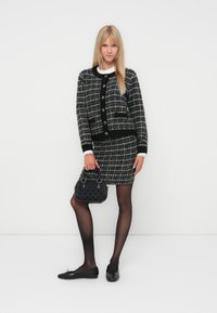 Ensemble de veste et jupe en tweed à carreaux noir et blanc, présentant un col rond, un bord noir contrastant, des poignets en dentelle et un sac à main texturé noir.