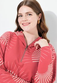 Haut à capuche de couleur rose avec un col zippé, présentant un motif de rayons de soleil en crème. Tissu doux avec des manches longues et une coupe décontractée.