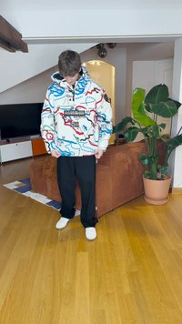 Weiße übergroße Kapuzenjacke mit einem bunten Kartenmuster, schwarzen Akzenten und Logo auf der Vorderseite. Gestylt mit lockeren schwarzen Hosen und weißen Turnschuhen.