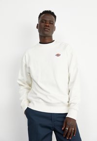 Sudadera blanca de algodón con puños y dobladillo acanalados, que presenta un pequeño logo azul y naranja en el pecho. El modelo lleva pantalones azul oscuro.