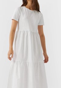 Femme portant une robe maxi blanche à manches courtes, à plusieurs niveaux, avec des coutures froncées et un col rond sur fond uni.