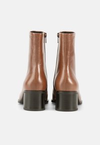 Ruskeat nahkaiset nilkkurit, joissa on sileä viimeistely, vetoketjusuljenta ja paksu musta korko. Design on tyylikäs ja minimalistinen.