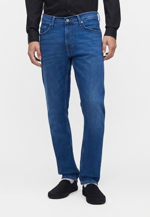 Person trägt gerade geschnittene blaue Jeans, schwarzes Langarmshirt, schwarze Slip-On-Schuhe und weiße Socken, steht auf weißem Hintergrund.