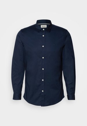 Camisa de botão azul-marinho feita de algodão, apresentando um logo discreto, mangas longas e um colarinho clássico. Botões brancos contrastam com o tecido.