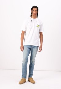 Hvid bomulds T-shirt med farverigt grafisk print på venstre bryst, parret med lyseblå denimjeans og brune sneakers.