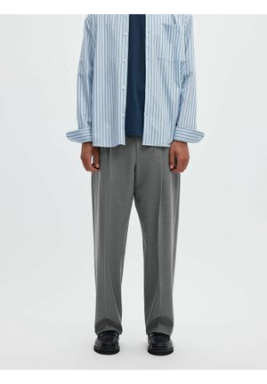 Personne portant un pantalon gris ample, des chaussures noires, une chemise bleu marine et une chemise rayée bleu clair déboutonnée à manches longues.