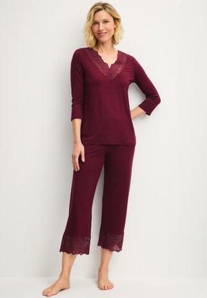 Donna in piedi a piedi nudi che indossa una maglia bordeaux a maniche lunghe e pantaloni corti con rifinitura in pizzo sullo scollo e sulle gambe dei pantaloni.