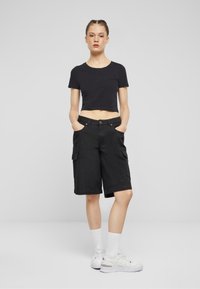 Kvinne står med hendene i lommene iført svart crop topp, svarte cargo shorts, hvite sokker og hvite joggesko mot en ensfarget bakgrunn.