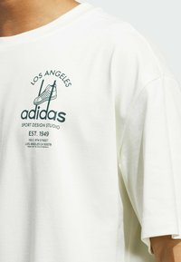 Baltas didelis marškinėliai su apvaliu iškirpimu. Yra žalias atspaudas su užrašais "Los Angeles," "adidas" ir batelio iliustracija.