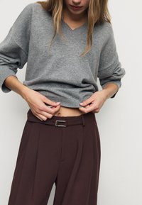 Grå V-ringad tröja med lösa ärmar, kombinerad med bruna högmidjade byxor med bälte och synliga pressveck.