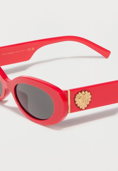 Κόκκινα οβάλ γυαλιά ηλίου με μαύρους φακούς, παχιά μπράτσα και χρυσό διακοσμητικό έμβλημα στο πλάι κοντά στη μεντεσέ.