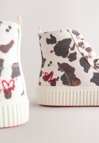 Aukštakulniai sportbačiai iš baltos ir rudos karvės rašto drobės; turi guminius pado, metalines akių skylutes ir raudonos širdies siuvinėjimą šone.