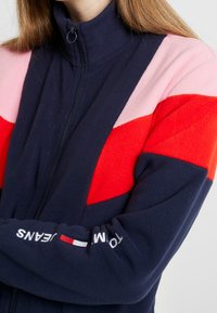 Fleecová bunda na zip v námořnické modři, červené, růžové a bílé barvě. Obsahuje design v barvách; vysoký límec a detaily loga na rukávu. Měkká textura.