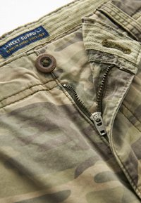 Kamuflerede cargo-short i dæmpede grønne og brune farver, med lynlås og knaplukning samt et vævet mærke på taljen.