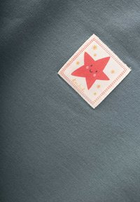 Červená usmievajúca sa hviezda s ružovými lícami a okolo nej malé žlté hviezdy na bielom štvorecovom poli na šedom látkovom pozadí.