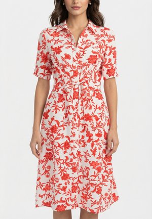 Femme portant une robe chemise midi blanche avec imprimé floral rouge, manches courtes, col et un nœud devant à la taille.