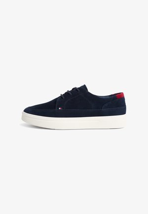 Marineblauwe suède sneaker met een witte rubberen zool, voorzien van een vetersluiting, gestikte accenten en een rood-wit detail op de hiel.